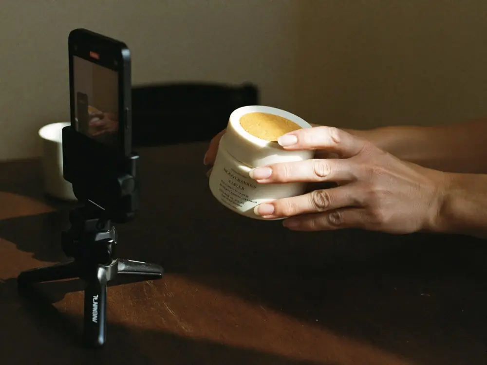 Symbolbild: Hand hält ein Produktglas vor ein Smartphone auf dem Stativ