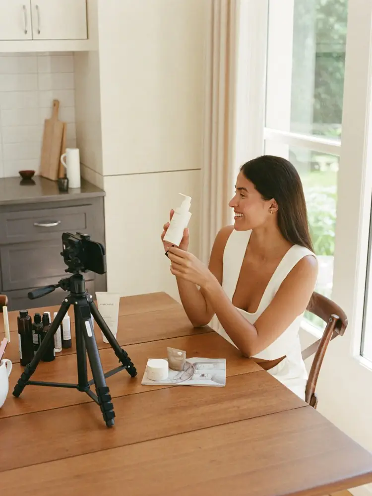 Symbolbild: Eine Frau hält am Küchentisch ein Produkt in die Smartphone-Kamera auf einem Stativ – so entsteht ein UGC-Video